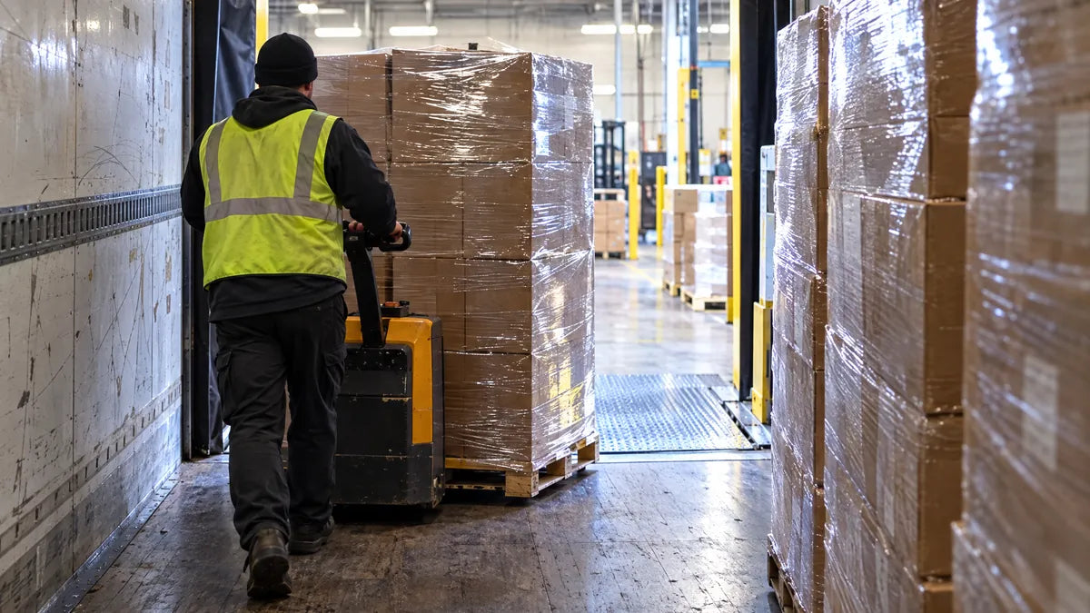 View from inside a trailer: a worker moves a stretch-wrapped pallet of cartons with a pallet jack